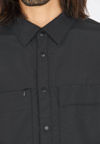 Zwarte blouse met een gestructureerde stof, voorzien van een puntige kraag, drukknoopsluitingen en een ritszak op de borst.