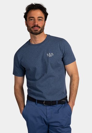 Man met donker haar en baard staat met handen in de zakken, draagt een blauw N.Z.A. New Zealand 1923 T-shirt en bijpassende blauwe broek met zwarte riem.