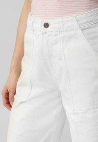 Białe dżinsowe spodnie z przednimi kieszeniami i metalowym zapięciem. Materiał jest gładki z widocznym szwem i luźnym krojem.