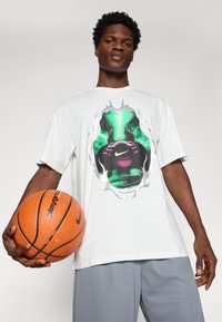 Hvid bomulds t-shirt med et grafisk tryk af en basketball med grønne accenter og en takkede ramme. Matchet med grå shorts og en basketball.