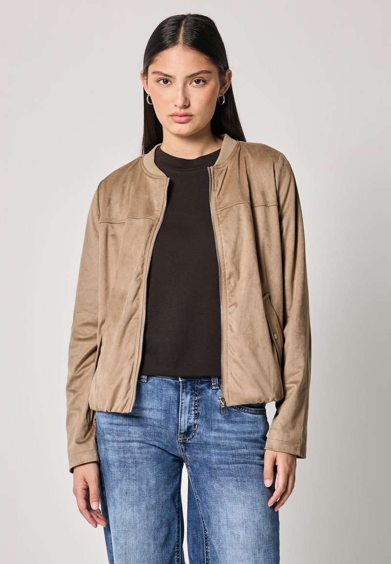 Giacca bomber in suede color crema con colletto rotondo, zip completa e tasche laterali, abbinata a una maglietta scura e jeans blu. Texture liscia.