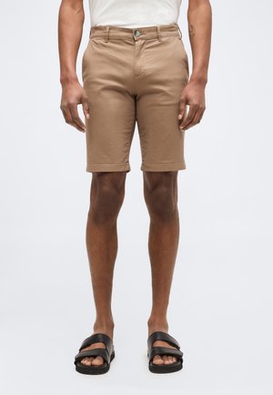 Homme portant un short beige jusqu’aux genoux, une chemise blanche et des sandales noires, debout devant un fond clair uni.