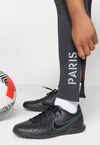 Fekete focicipők érdes felülettel és teal színű Nike logóval, szürke, tapered fazonú nadrággal, amelynek oldalán a "PARIS" felirat szerepel. Egy focilabda van a közelben.