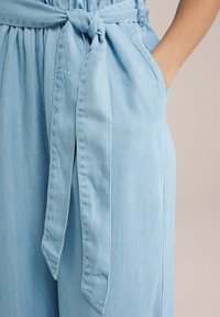 Lyseblå denim jumpsuit med en bundet talje-sash, sidelommer og en afslappet pasform. Har subtil vertikal tekstur og pæne syninger.
