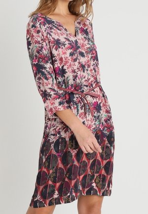 Kvinde iført knælang blomstrede kjole med 3/4 ærmer og et tyndt bælte, der kombinerer rosa, lilla, sort og grønne toner med cirkulære mønstre.