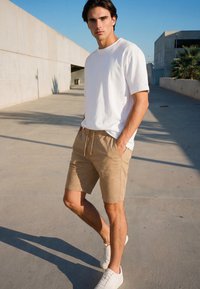 Young man standing outdoors in sunlight wearing a white t-shirt, beige shorts, and white sneakers with hands in pockets.
