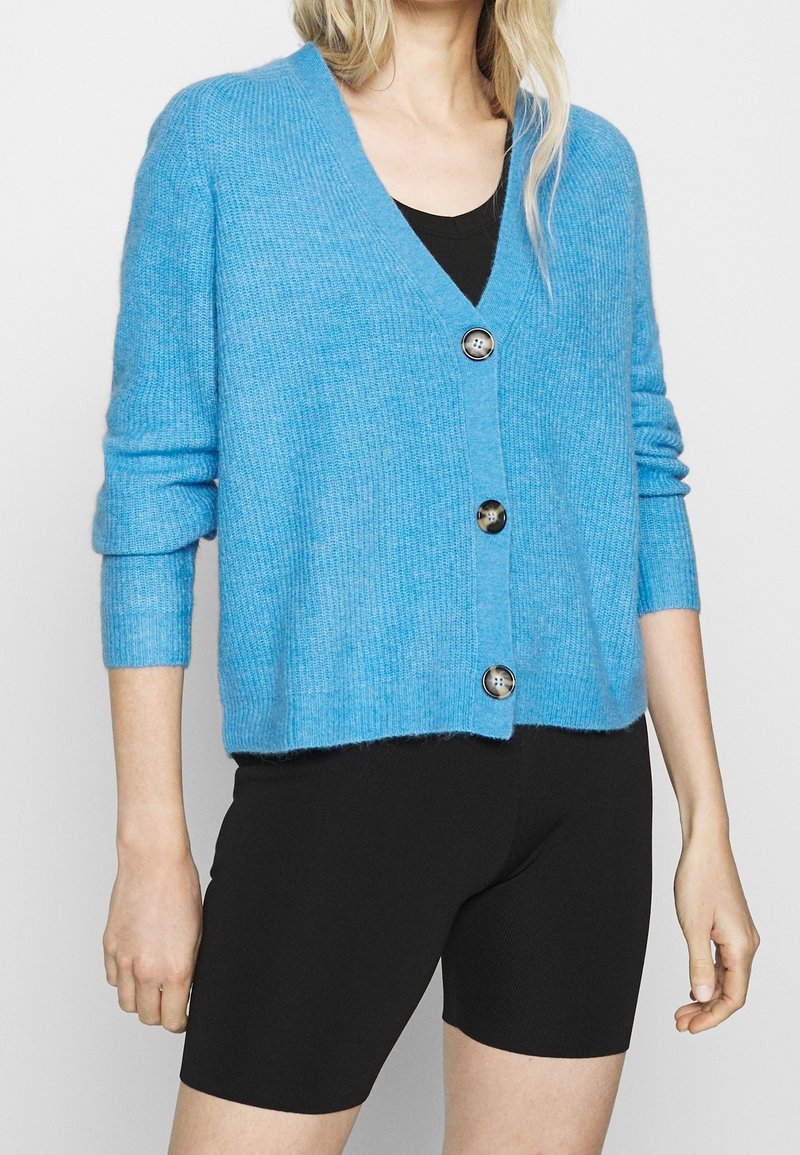 Femme portant un cardigan bleu clair boutonné sur un top noir en V et un short noir ajusté sur un fond uni.