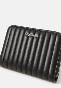 Čierna kožená peňaženka s prešívaným dizajnom, s vertikálnym vystužením. Strieborné logo „Calvin Klein“ a zips. Kompaktný obdĺžnikový tvar.