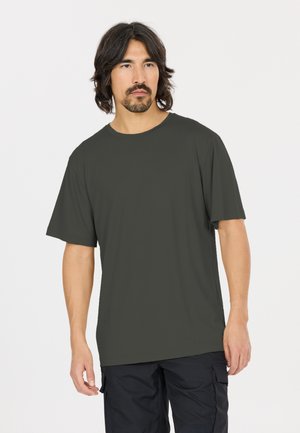 Homme aux cheveux mi-longs et barbe foncés, portant une chemise à manches courtes gris foncé et un pantalon cargo noir, debout devant un fond blanc.