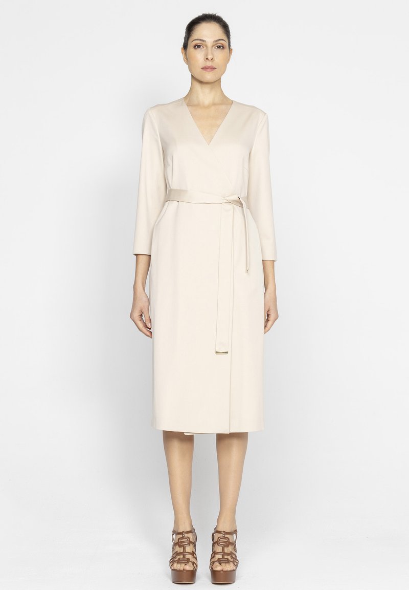 Beżowa sukienka typu wrap z rękawami 3/4; posiada pasek do przewiązania na talii oraz długość do kolan. Gładka tekstura materiału.