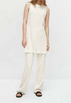 Kobieta w białej dzianinowej tunice bez rękawów i dopasowanych szerokich spodniach dzianinowych, nosząca czarne sandały z odkrytymi palcami, na jednolitym tle.