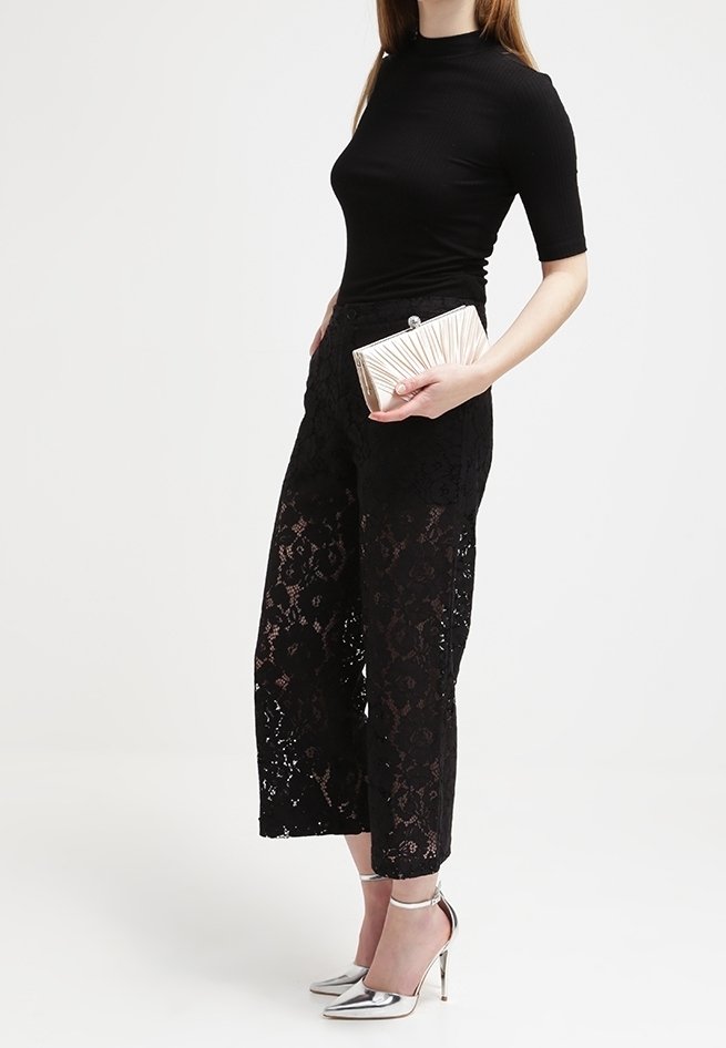 Kobieta w czarnych koronkowych spodniach szerokich nogawkach, dopasowanej czarnej bluzce, srebrnych szpilkach, trzymająca złotą torebkę teksturowaną na białym tle.