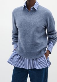 Personne portant un pull bleu col en V par-dessus une chemise à rayures bleues et blanches, les mains dans les poches d’un jean bleu foncé.