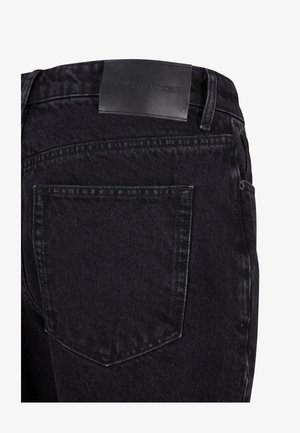 Mustat farkut, joissa on teksturoitu pinta, ommeltuja yksityiskohtia ja vyötäröllä nahkatarra, jossa on kohopainatus. Yksi takatasku.