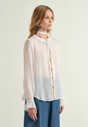 Femme aux longs cheveux blonds portant un chemisier blanc transparent boutonné avec un col noué d'un ruban et un pantalon bleu à larges jambes, debout devant un fond uni.