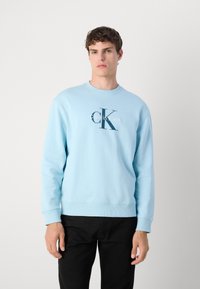 Világoskék pulóver bordás mandzsettával és aljjal, amelyen egy nagy sötétkék "CK" logó és a "Calvin Klein" felirat látható az elején.