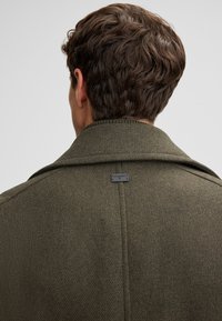 Manteau en laine vert olive avec une finition texturée, grand col et détail de tag au dos. Présente une silhouette ajustée et des accents de couture visibles.