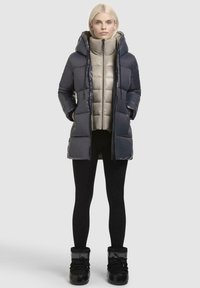 Gevoerde jas met een donkergrijze buitenlaag, lichtgrijze binnenlaag, hoge kraag, capuchon en ritssluiting. Gemaakt met zwarte leggings en laarzen.