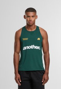 Muž stojící v tmavě zeleném bezrukávovém sportovním tričku s nápisem "another" vytištěným bílou barvou na hrudi a v černých šortkách.