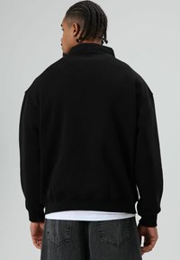 Zwarte trui met hoge kraag, losse pasvorm en geribbelde manchetten. Draagt over een wit overhemd en gecombineerd met grijze jeans. Eenvoudig ontwerp.