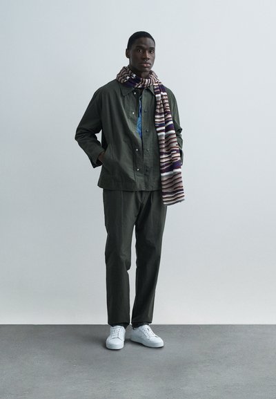 Πράσινο μπουφάν και παντελόνι με μπλε κουμπιά, συνδυασμένα με πολύχρωμο πλεκτό κασκόλ και λευκά αθλητικά παπούτσια, παρουσιάζονται σε ουδέτερο φόντο.