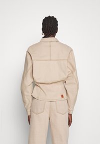 Veste en denim beige au design ajusté, avec des coutures marquées et un ourlet légèrement arrondi. Elle présente une étiquette dans le bas du dos.