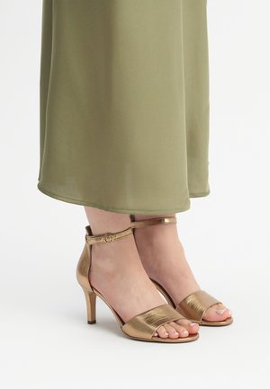 Bronze metallic high-heeled sandals with a wide toe strap and an ankle strap featuring a round buckle. Worn with a green flowy dress.