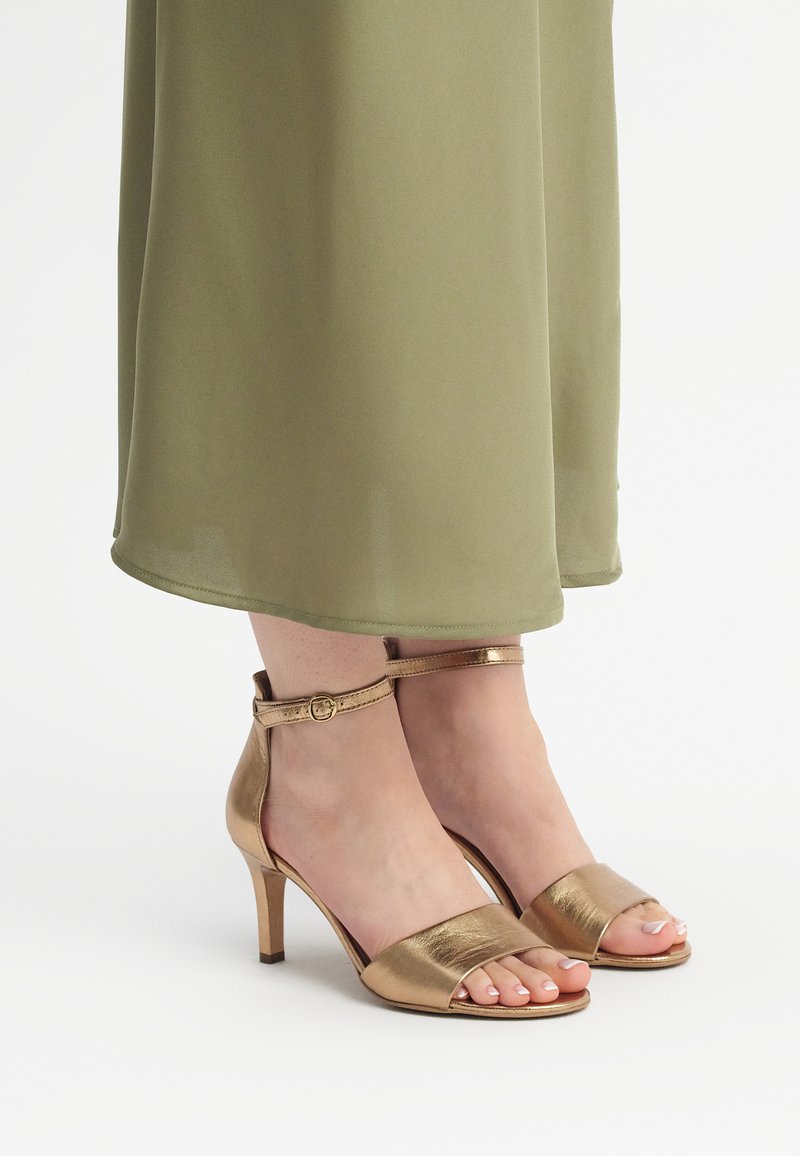 Bronze metallic high-heeled sandals with a wide toe strap and an ankle strap featuring a round buckle. Worn with a green flowy dress.