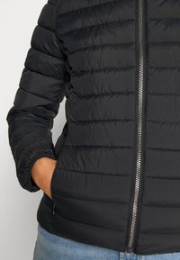 Personne portant une veste matelassée noire avec une fermeture éclair argentée, main gauche dans la poche, associée à un jean en denim bleu.