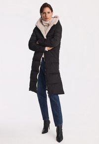 Czarny, pikowany długi płaszcz z jasnym futrzanym kapturem i podszewką, zapinany na zamek, noszony na białym swetrze i niebieskich dżinsach. Czarne botki na obcasie.