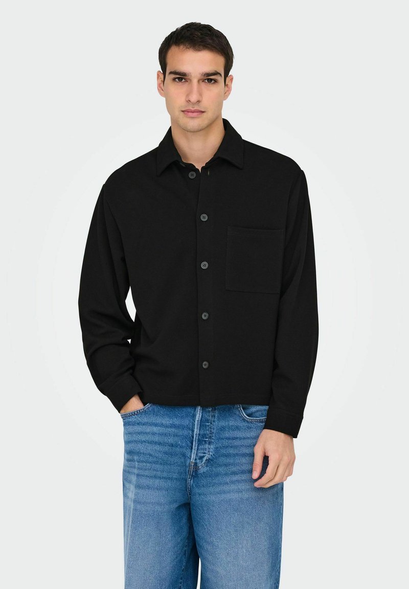 Black button-up shirt with a collar, long sleeves, and a chest pocket. Made of solid fabric, paired with light blue jeans.
