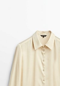 Blouse beige à manches longues en tissu lisse. Présente un col classique, un devant à boutons et un design minimaliste sans motifs.