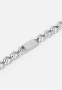 Zilverkleurige kettingarmband met ovale schakels en een rechthoekige sluiting met gegraveerd "CK" logo. Gladde, metallic afwerking.