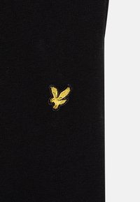 Melnā audumā ar gludu tekstūru ir neliels dzeltenais izšūtais putna logo augšējā daļā, piedāvājot kontrastējošu detaļu.