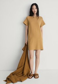 Rochie tricotată, cu mânecă scurtă, de culoare muștar, cu o croială lejeră, purtată cu un șal texturat în nuanța asemănătoare. Sandale maro completează ținuta.