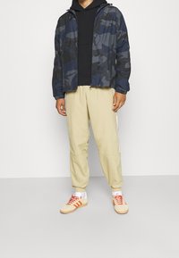 Marinblå kamouflageskjorta, svart hoodie, beige joggers med vita ränder och beige sneakers med orange detaljer. Avslappnad outfit med blandade texturer.