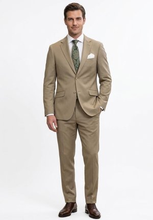 Hombre con traje color beige, corbata verde con diseño, camisa blanca, pañuelo blanco en el bolsillo y zapatos marrones, de pie con una mano en el bolsillo.