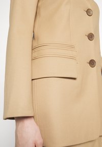 Blazer beige avec un design structuré, trois grands boutons marron et des coutures détaillées sur les poches. Tissu lisse et texturé.