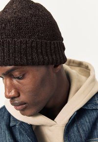 Cappello a cuffia marrone lavorato a maglia con risvolto a costine e superficie testurizzata. Indossato con una felpa beige con cappuccio e una giacca di jeans, mostra il profilo laterale del modello.