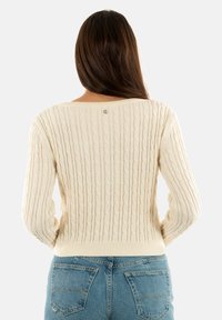 Suéter de punto color crema con un patrón de trenzas, cuello redondo y puños acanalados. Presenta un pequeño logo en la parte posterior. Lleva encima de unos jeans azules.