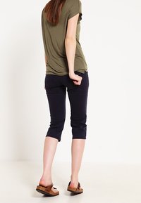 Oliwkowa bluzka z krótkim rękawem, luźny krój, połączona z granatowymi spodniami 7/8. Modelka nosi brązowe sandały na slip-on, prezentując casualowy strój.