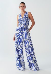 Plavi i bijeli kombinezon s uzorkom, omotnog dizajna, širokih nogavica i vezicom u struku. Izrađen od lagane tkanine glatke teksture.