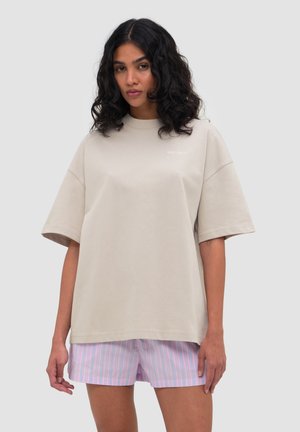 Žena s tmavými kudrnatými vlasy, která má na sobě oversized béžové tričko a pastelově růžové a modré pruhované šortky na jednobarevném pozadí.