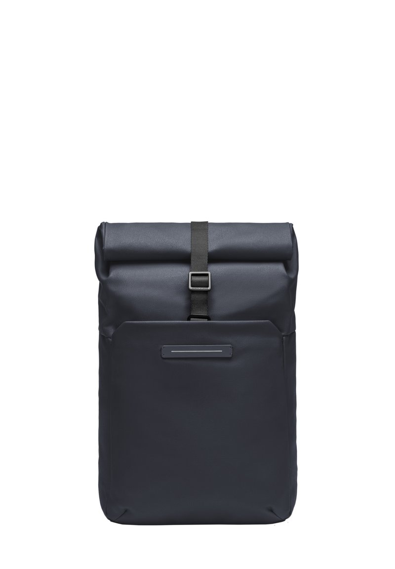 Sac à dos à rabat en tissu bleu foncé, avec une poche avant, une fermeture par boucle en métal et une surface texturée. Design compact pour une utilisation urbaine.