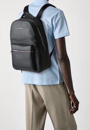 Personne portant une chemise bleu clair à manches courtes et un pantalon beige, portant un sac à dos en cuir noir texturé avec une poche frontale zippée.