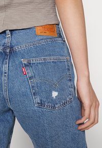 Blå denimjeans med en blek tvätt, ett synligt hål och en röd Levi's-flik på bakfickan. Har en brun läderetikett på midjebandet.