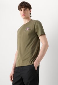 Camiseta de algodón verde oliva con cuello redondo, mangas cortas y logo bordado que presenta un escudo y texto en el pecho.