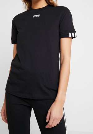 Vrouw draagt een zwart Adidas T-shirt met een klein logoplaatje op de borst en drie witte strepen op de mouwboorden, gecombineerd met zwarte broek.