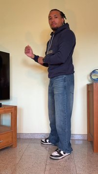 Moleton azul-marinho com bolso frontal, jeans de ganga azuis e ténis pretos e brancos. Em pé perto de um móvel de madeira e da televisão.