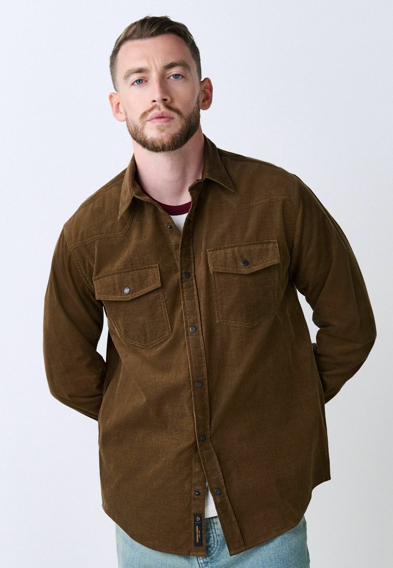 Brown corduroy shirt with a button-down collar, two chest pockets, and dark buttons. Features a textured fabric and relaxed fit.